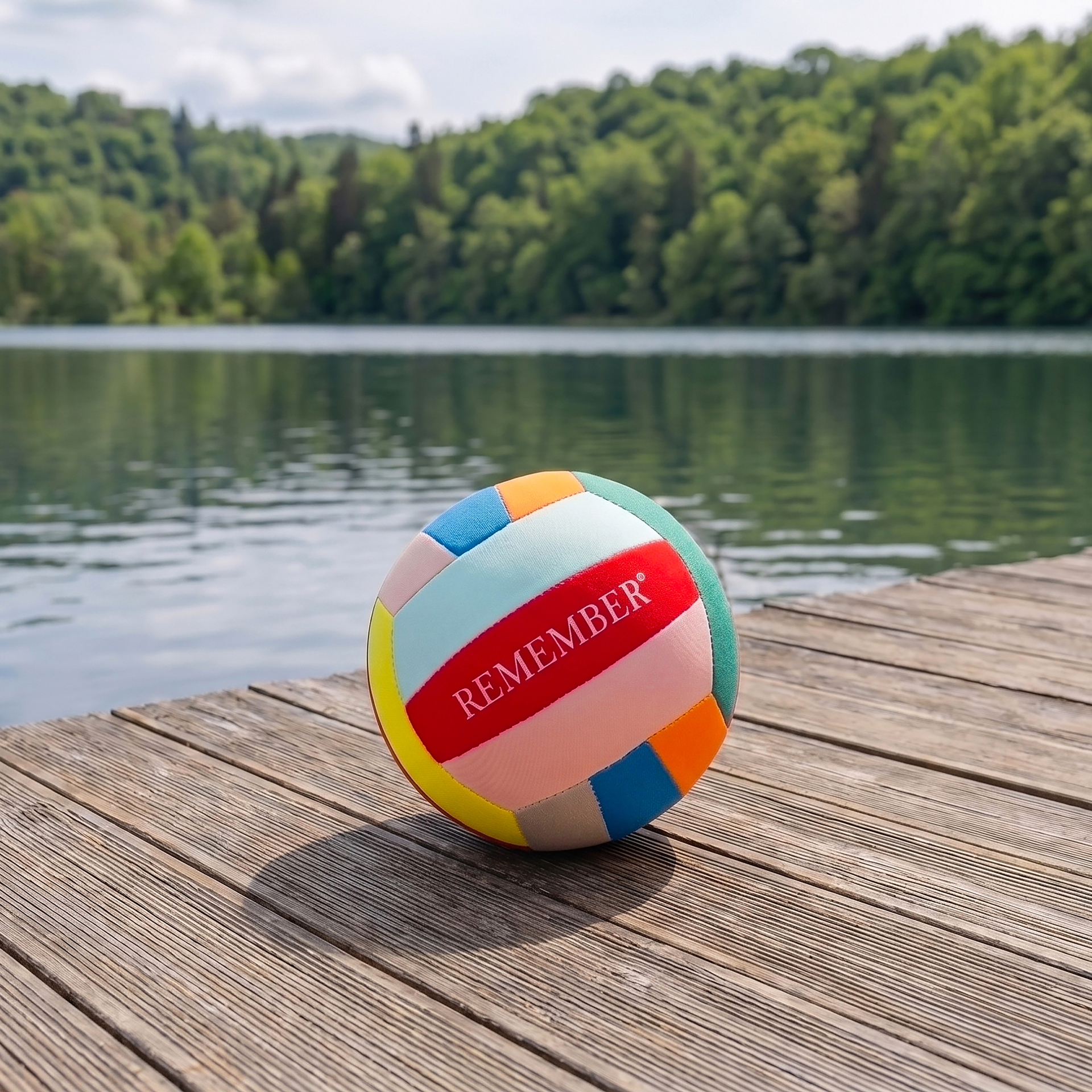 A colourful ball, similar to a beach or volleyball, with the word ‘ REMEMBER ’ in red on a light pink background, lies on a sun-drenched wooden jetty. Behind it lies a tranquil lake, surrounded by green forests beneath a slightly cloudy sky.