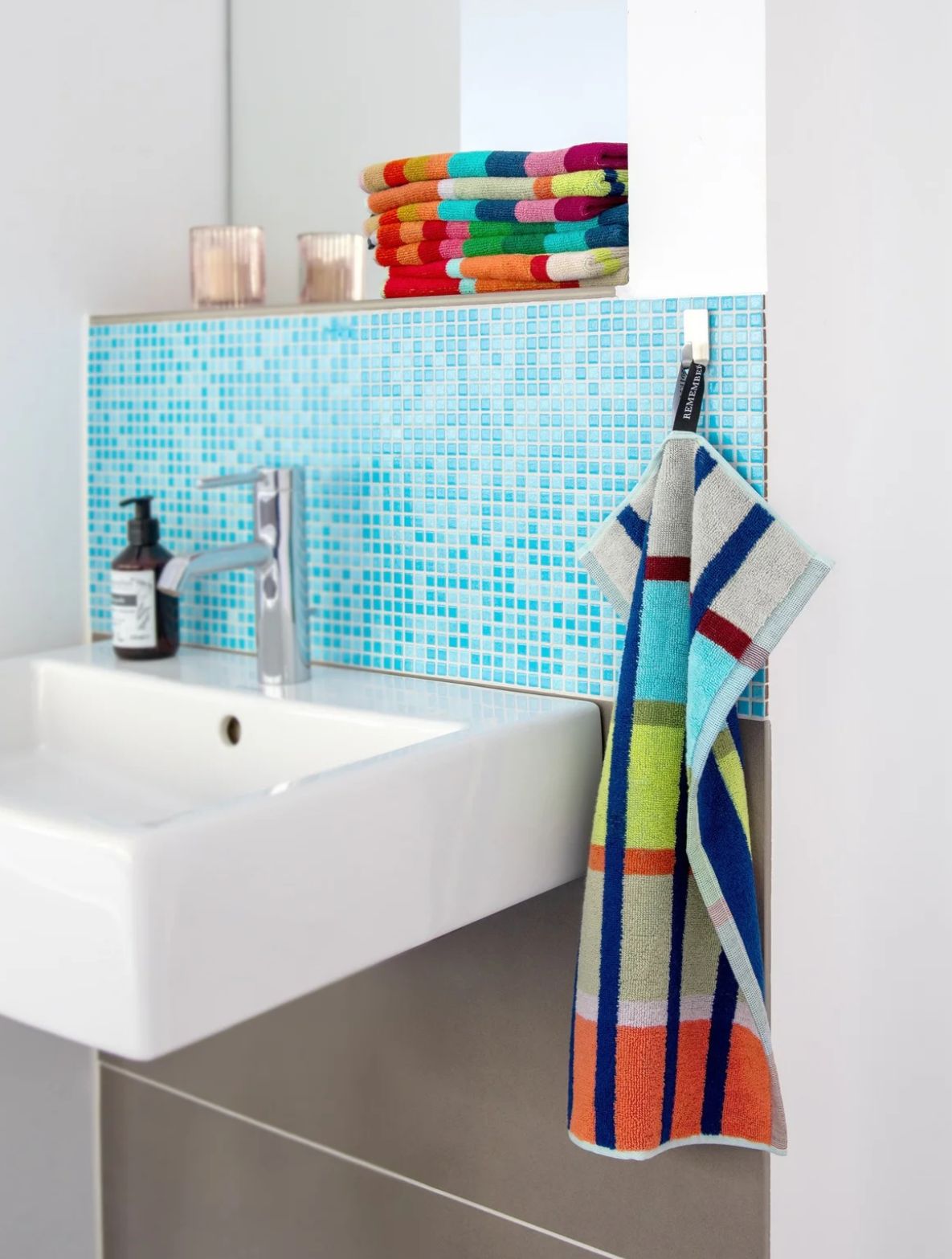 Modernes Badezimmer mit weißem Waschbecken, verchromtem Wasserhahn und braunem Seifenspender. Dahinter blaue Mosaikfliesen. Auf einem Regal liegen bunte, gestreifte Handtücher und zwei Kerzenhalter. Rechts hängt ein Handtuch.