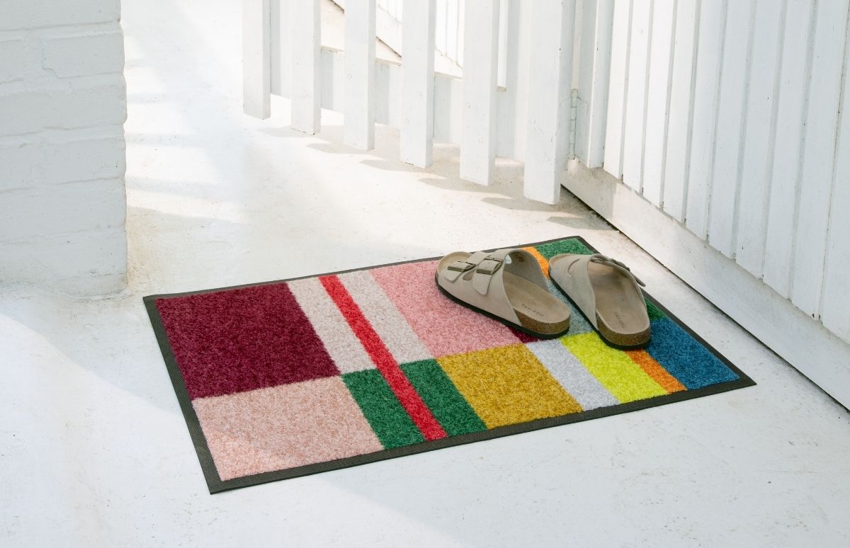 Eine bunte, rechteckige Fußmatte mit geometrischem Muster in Dunkelrot, Rosa, Grün, Gelb, Orange und Blau liegt auf hellem Boden. Darauf stehen zwei helle, offene Sandalen mit Schnallen. Im Hintergrund: weißer Holzzaun, Sonnenlicht.