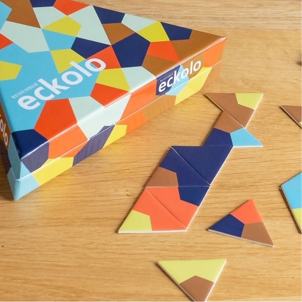 Bunter, dreieckiger Spielekarton von „eckolo“ auf Holztisch, mit geometrischem Muster aus blauen, orangen, gelben und braunen Polygonen. Daneben dreieckige Puzzleteile und eine Zickzack-Reihe im gleichen Design.