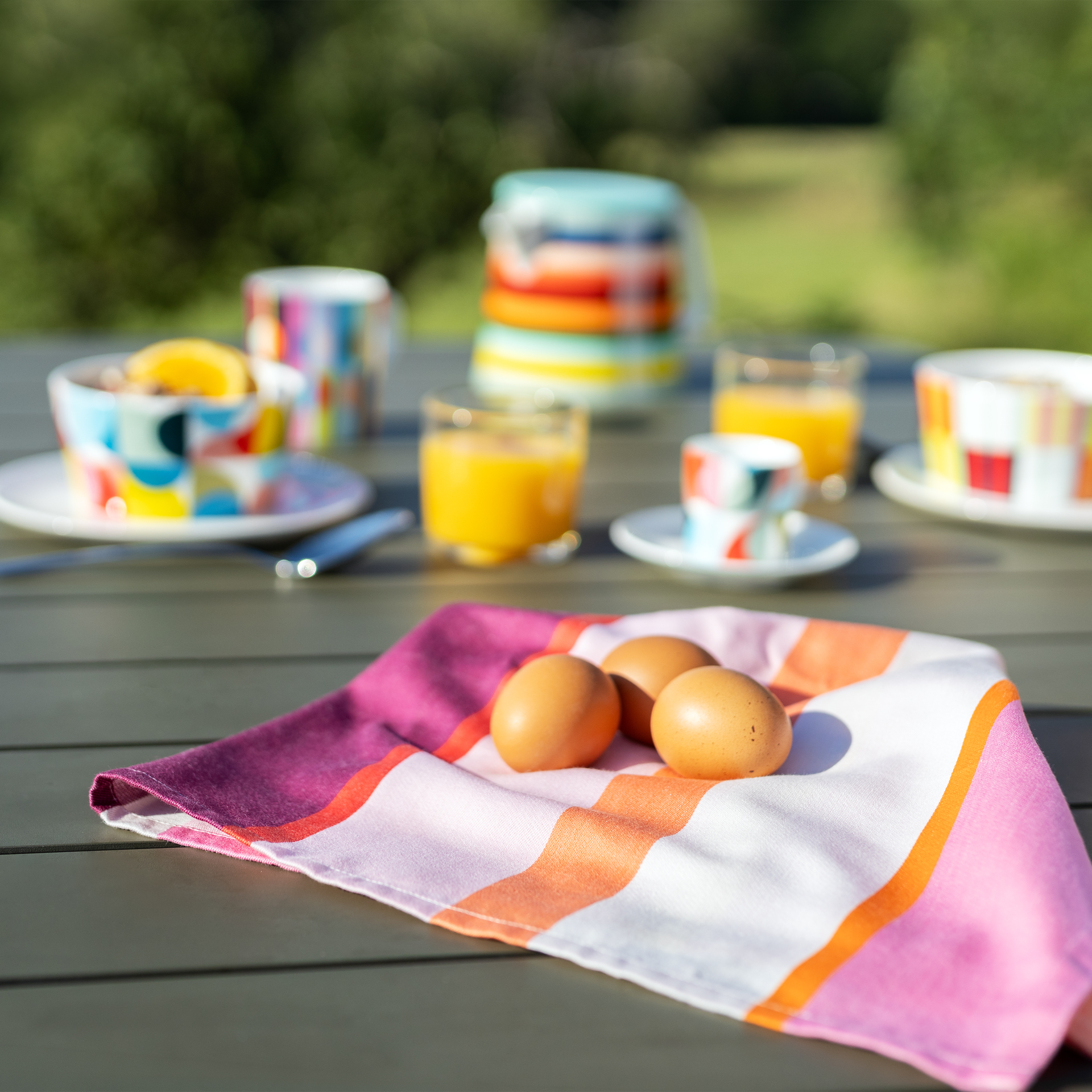 Sobre una mesa de madera oscura hay un colorido paño de cocina con rayas anchas en rosa, naranja, morado y blanco. Sobre él descansan tres huevos marrones. Al fondo, difuminado, se ven vajilla de colores para el desayuno y zumo de naranja al aire libre.