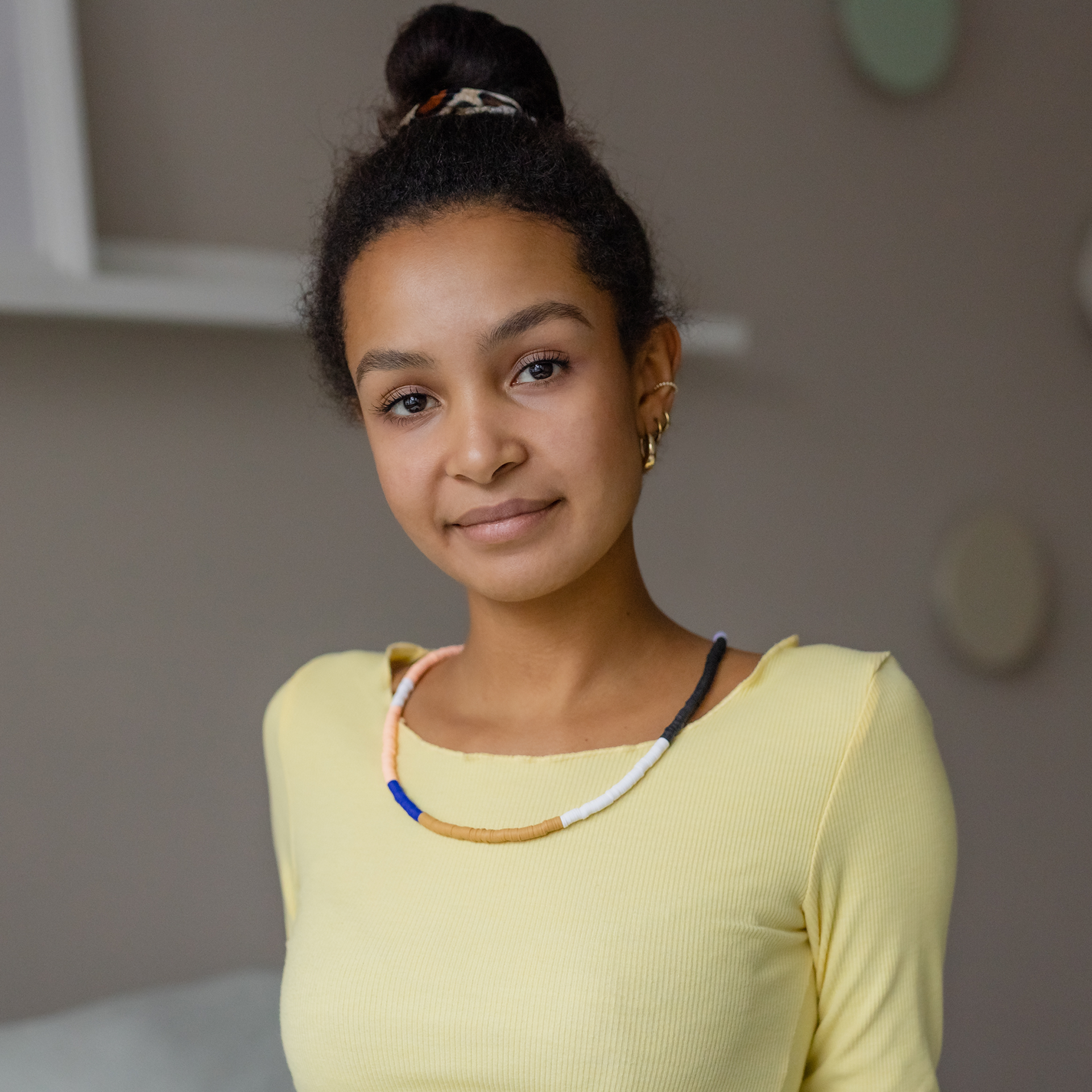 Jonge vrouw met donkere huidskleur en haar in een knot kijkt vriendelijk in de camera. Ze draagt een pastelkleurige parelketting (roze, blauw, beige, wit, donkergrijs) over een lichtgele blouse. Gouden oorbellen.