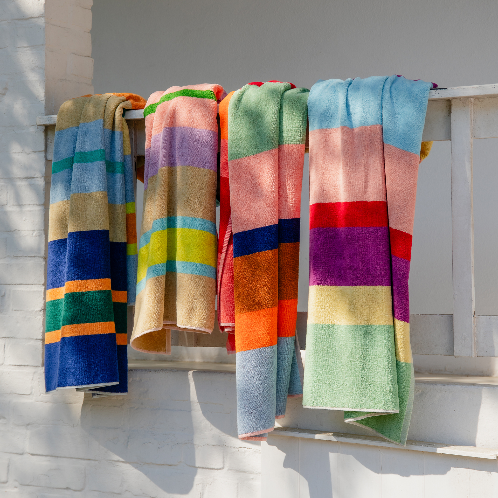 Serviettes de bain moelleuses à rayures colorées suspendues à une balustrade blanche, au soleil. Grandes serviettes en coton aux combinaisons de couleurs originales: bleu, vert, orange, beige, rose, rouge, violet et jaune.