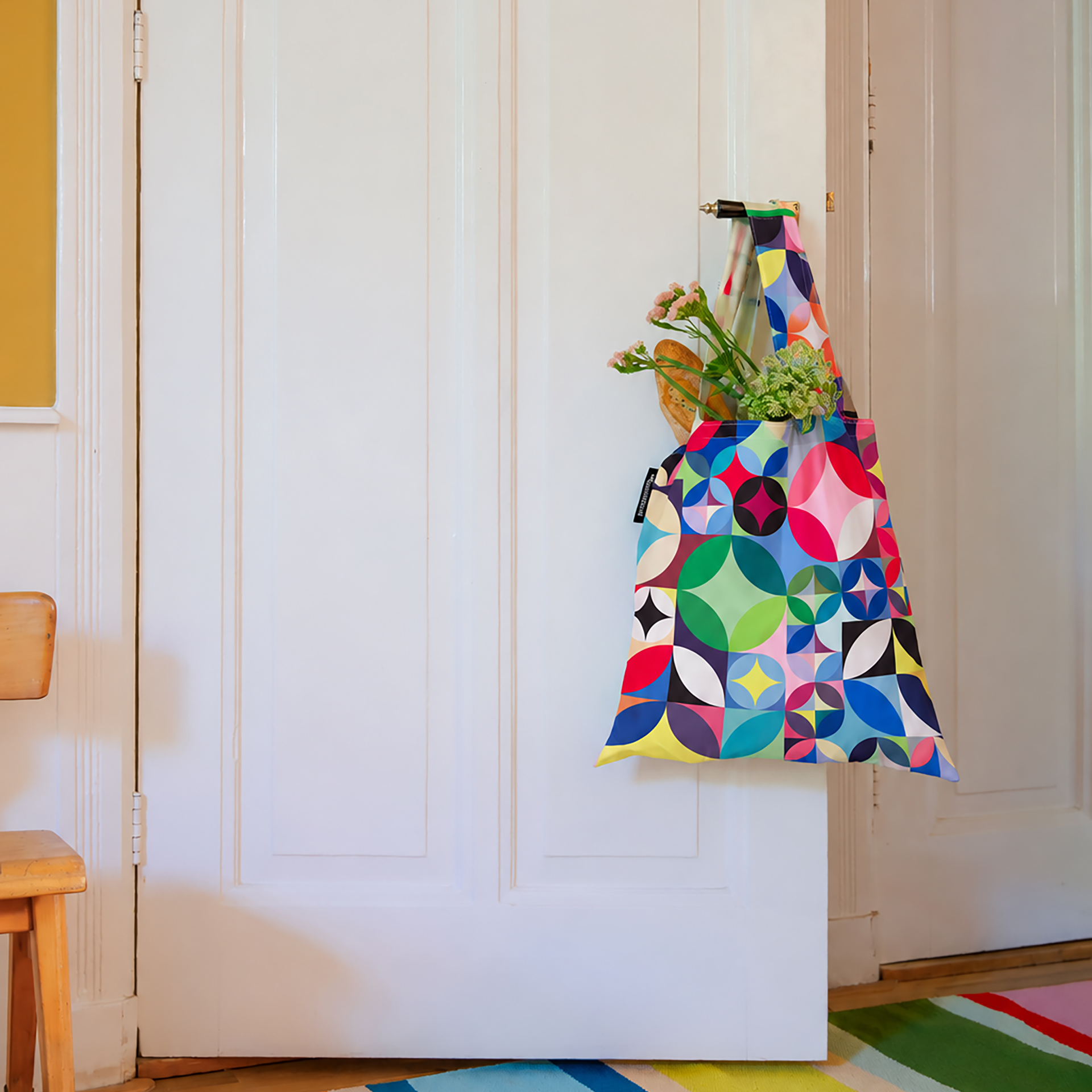 Eine bunte Tragetasche mit geometrischem Rosetta-Muster hängt an einer weißen Zimmertür. In der Stofftasche sind ein Baguette und frische rosa-grüne Blumen. Die Tür ist leicht geöffnet, ein gestreifter Teppich liegt auf Holzboden.