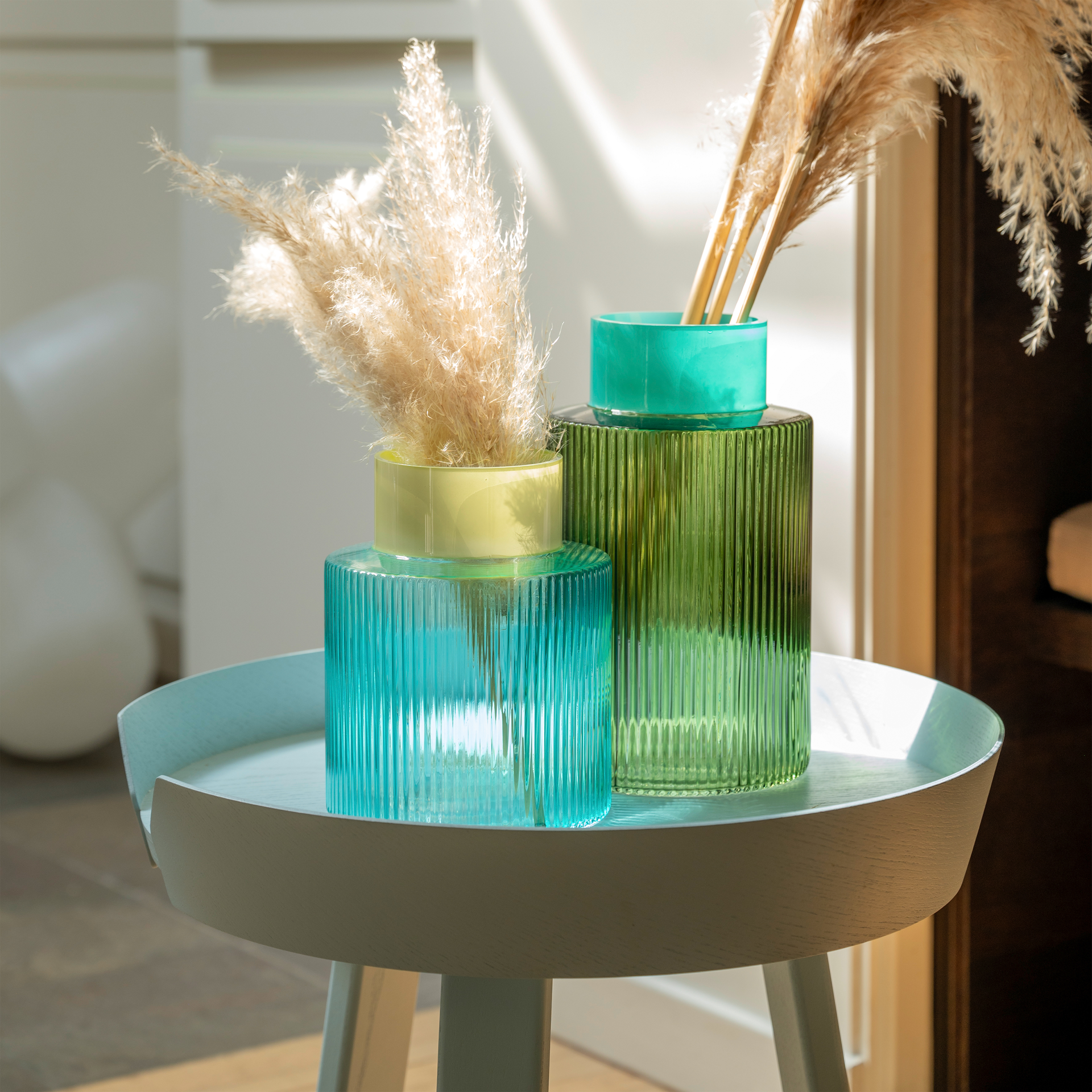 A blue and a green glass vase with grooves stand on a table and are decorated with decorative grass.