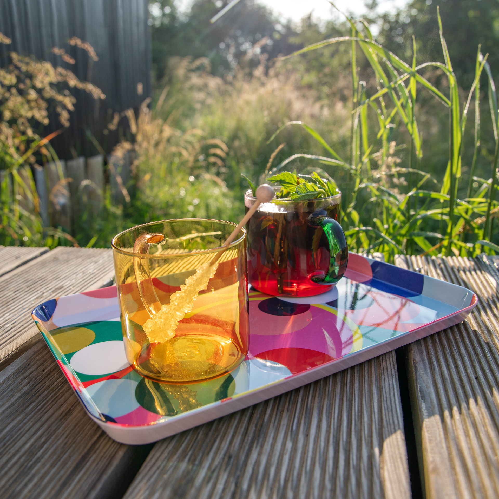 Dos vasos de cristal «Fred» sobre una bandeja de colores vivos en una terraza de madera. En el vaso de color rosa-amarillo hay una bebida clara con azúcar cande, y en el rojo con asa verde, una oscura con menta. Detrás, hierba difuminada a la luz del sol.