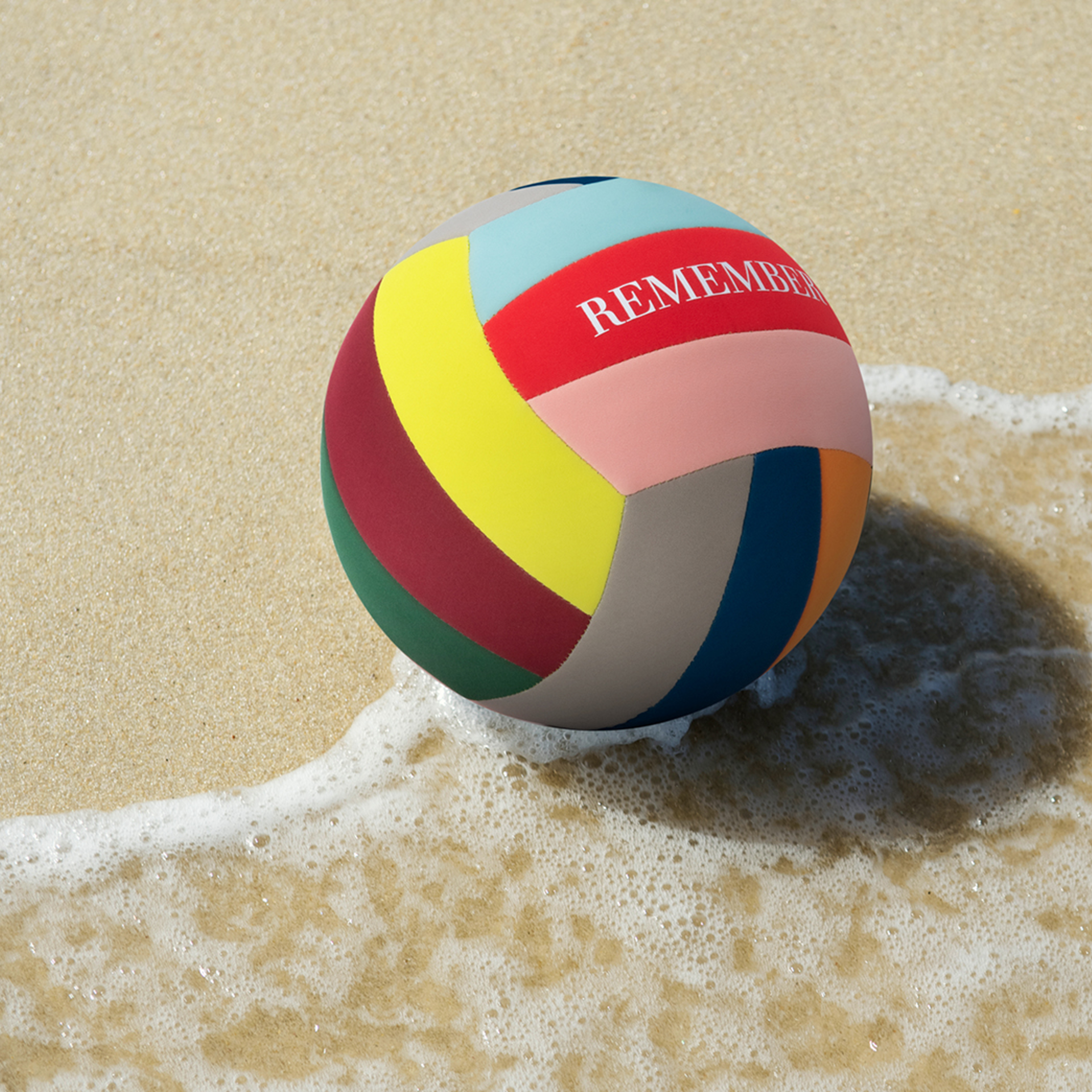 A colourful leisure ball lies on the beach, partly in the bright water. The words "REMEMBER" are written on a red segment.