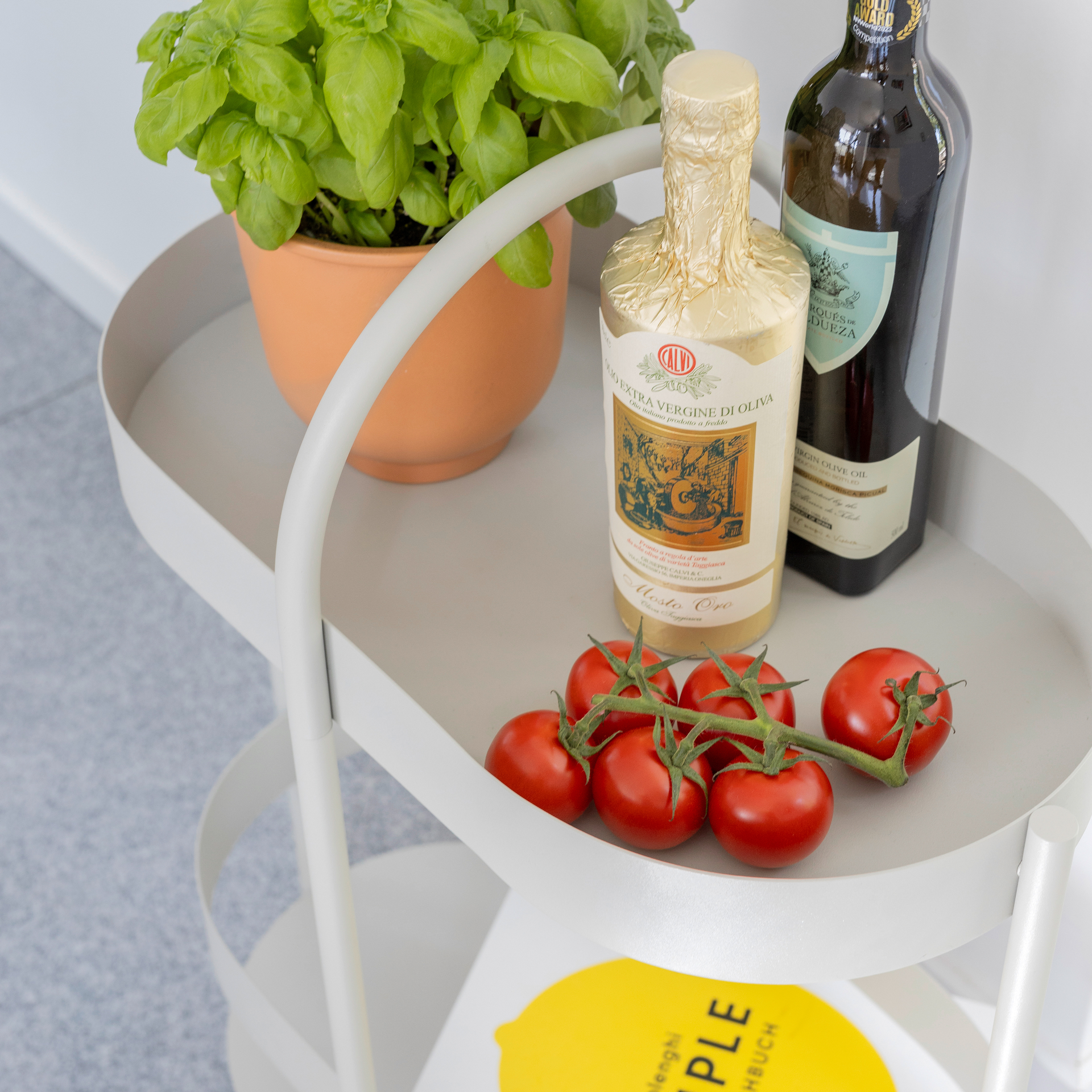 La consola auxiliar clara Barbara con albahaca, aceite de oliva, tomates y un libro. Se aprecian dos niveles y un tirador.