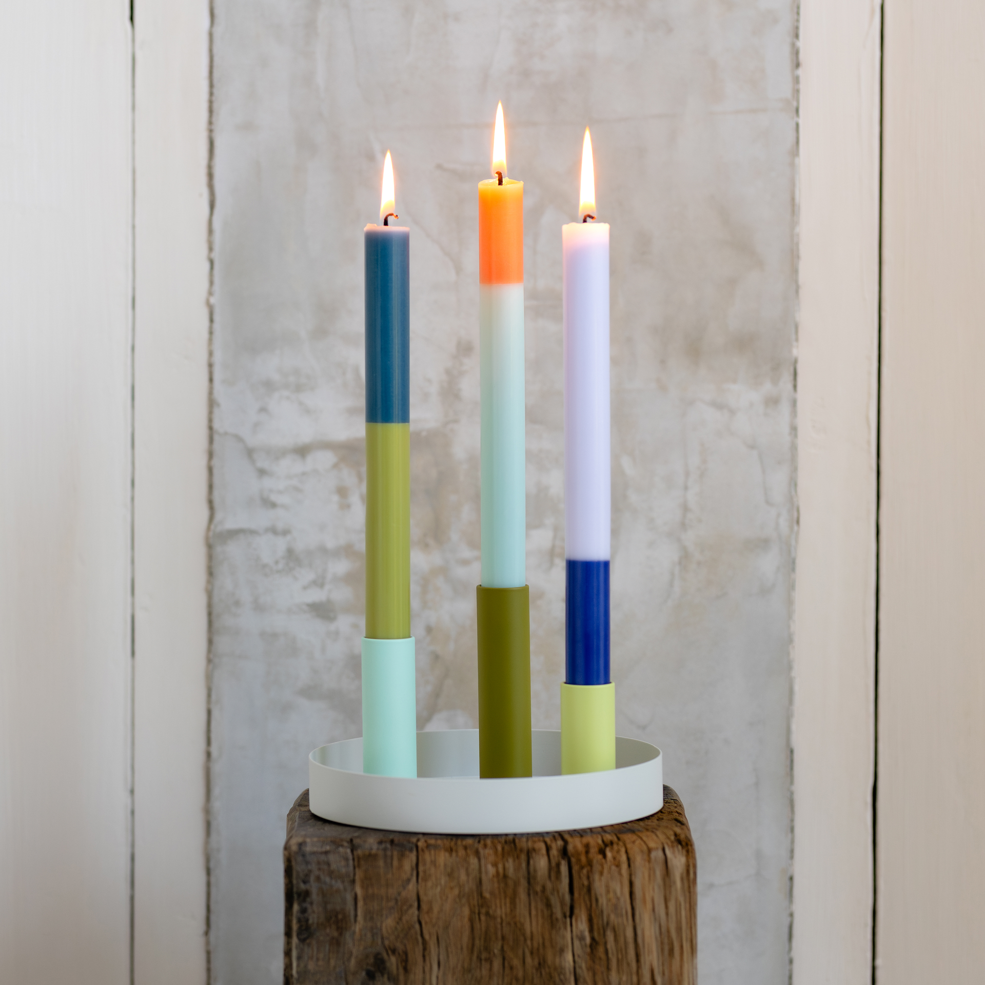 Tres velas cilíndricas de colores, en portavelas magnéticos, descansan sobre una bandeja blanca. Las velas están encendidas. El juego de portavelas «Luna Mini» descansa sobre un taburete de madera.