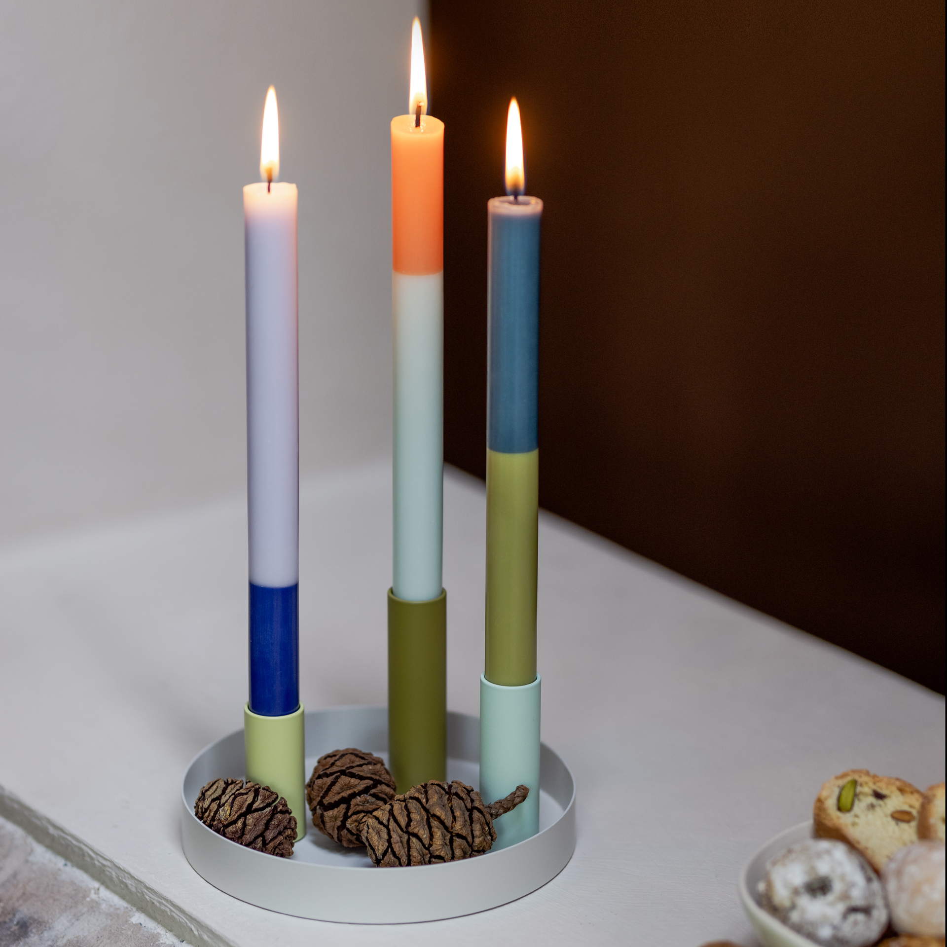 Tres velas cilíndricas de colores «Riga» encendidas en un candelabro. Las velas son de color blanco/azul, blanco/naranja y verde/azul. En primer plano hay piñas y galletas.