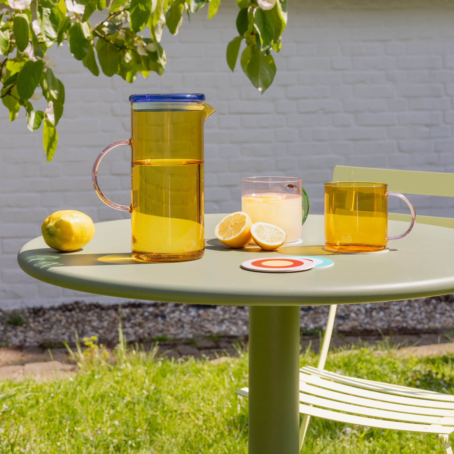 Ein bernsteinfarbener Glaskrug mit gelbem Getränk, zwei Gläsern, Zitronen und bunten Untersetzern auf einem hellgrünen Gartentisch. Im Hintergrund blühende Zweige, grüne Blätter und eine weiße Mauer. Eine frische, sommerliche Atmosphäre.