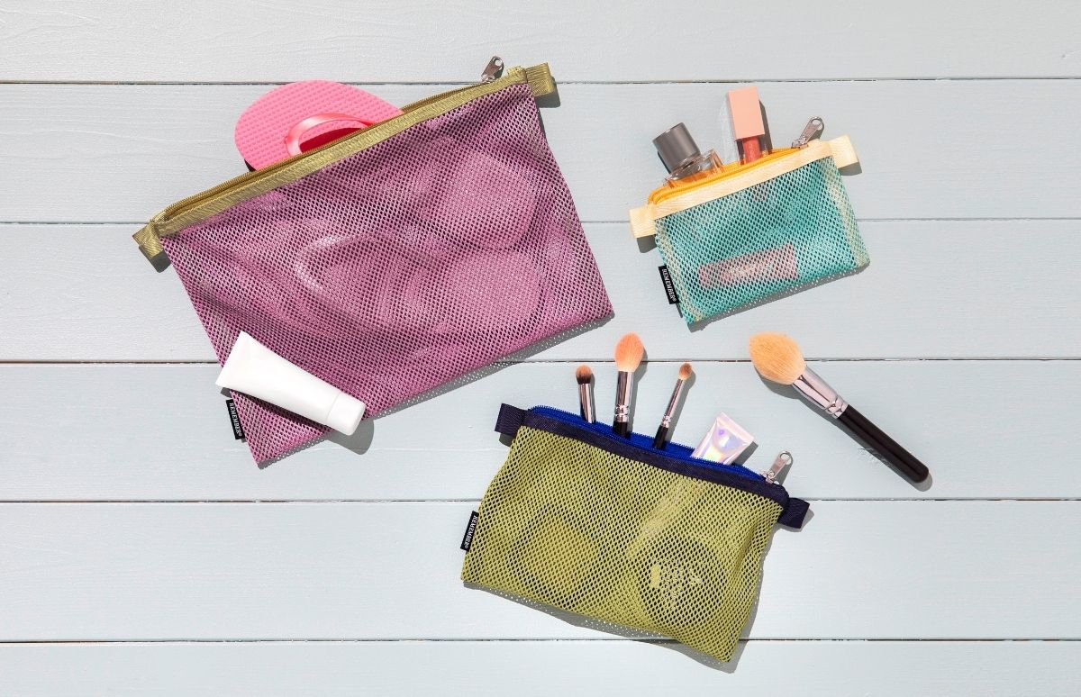 Drei bunte Netztaschen auf hellem Holzboden. Die pinke Tasche enthält Flip-Flops und Wattepads, daneben eine weiße Sonnencreme-Tube. Türkis birgt Parfüm und Lipgloss. Limonengrün: Pinsel und holografische Tube.