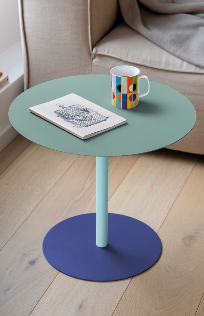 Runder Beistelltisch mit mintgrüner Platte, hellblauem Ständer und dunkelblauem Fuß auf hellem Holzboden. Darauf: geöffnetes Notizbuch mit Vogelkäfig-Skizze und bunte Tasse mit Streifen- und Geomuster. Beige Couch im Hintergrund.