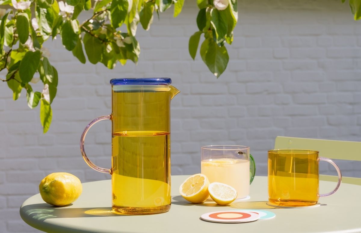 Sonniger Sommertag: Auf hellgrünem Gartentisch steht eine hohe gelbe Glaskaraffe mit blauem Deckel und erfrischendem Getränk. Daneben eine Zitrone und zwei halbe. Glas Limonade, leeres gelbes Glas. Blühende Äste, weiße Ziegelwand.
