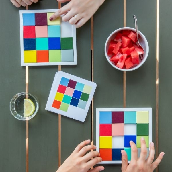 Zwei Personen lösen Farbpuzzles auf einem dunkelgrünen Holztisch mit kupferfarbenen Streifen. Eine Hand schiebt einen hellgelben Stein oben, während unten zwei Hände arbeiten. Daneben: Wassermelone, Zitronenwasser, Farbkarte.