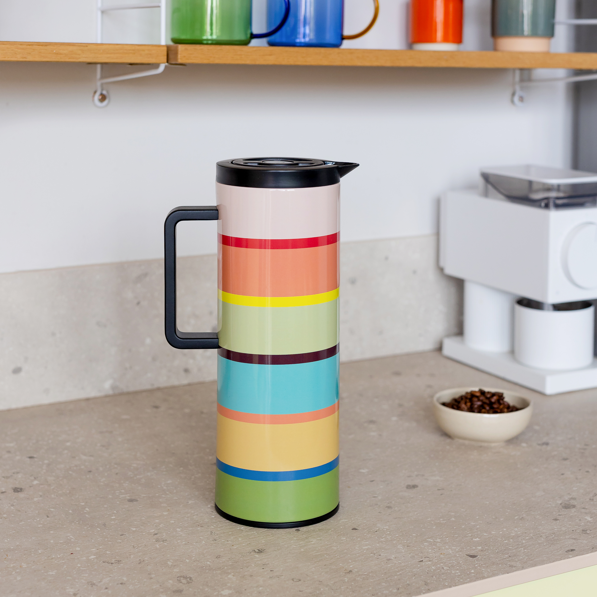 Colourful vacuum flask in a striped design with horizontal pastel stripes (beige, red, orange, yellow, light green, dark brown, turquoise, light blue, green). Black lid with spout and sturdy black handle. Set against a white background.