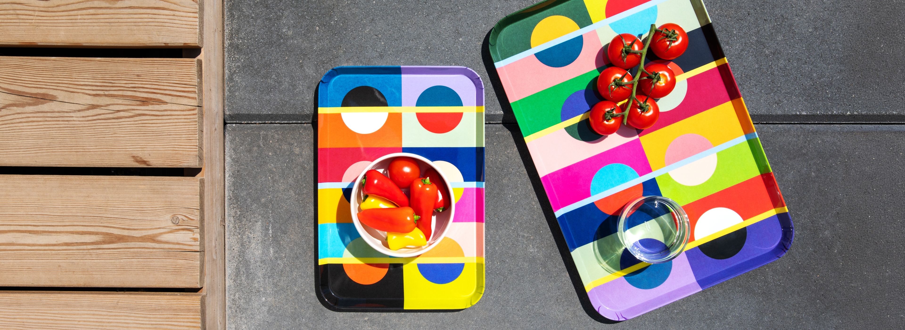 Zwei bunte Tabletts mit geometrischen Mustern stehen auf dunklem Beton neben einer Holzterrasse. Links: weiße Schale mit roten Kirschtomaten sowie gelben und roten Mini-Paprika. Rechts: Bund Kirschtomaten und Trinkglas.