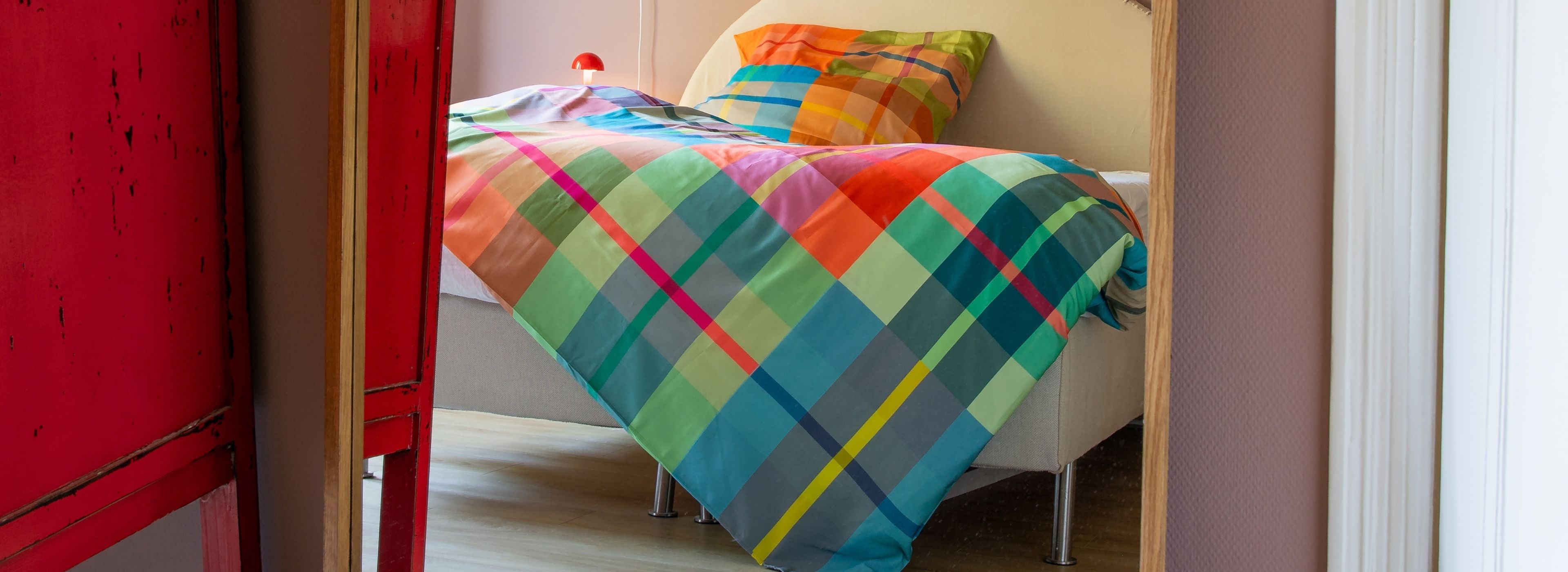 Bunt kariertes Bett mit Decke und Kissen in hellem Zimmer. Links rotes, abgenutztes Möbel; hinten kleine rote Pilzlampe.