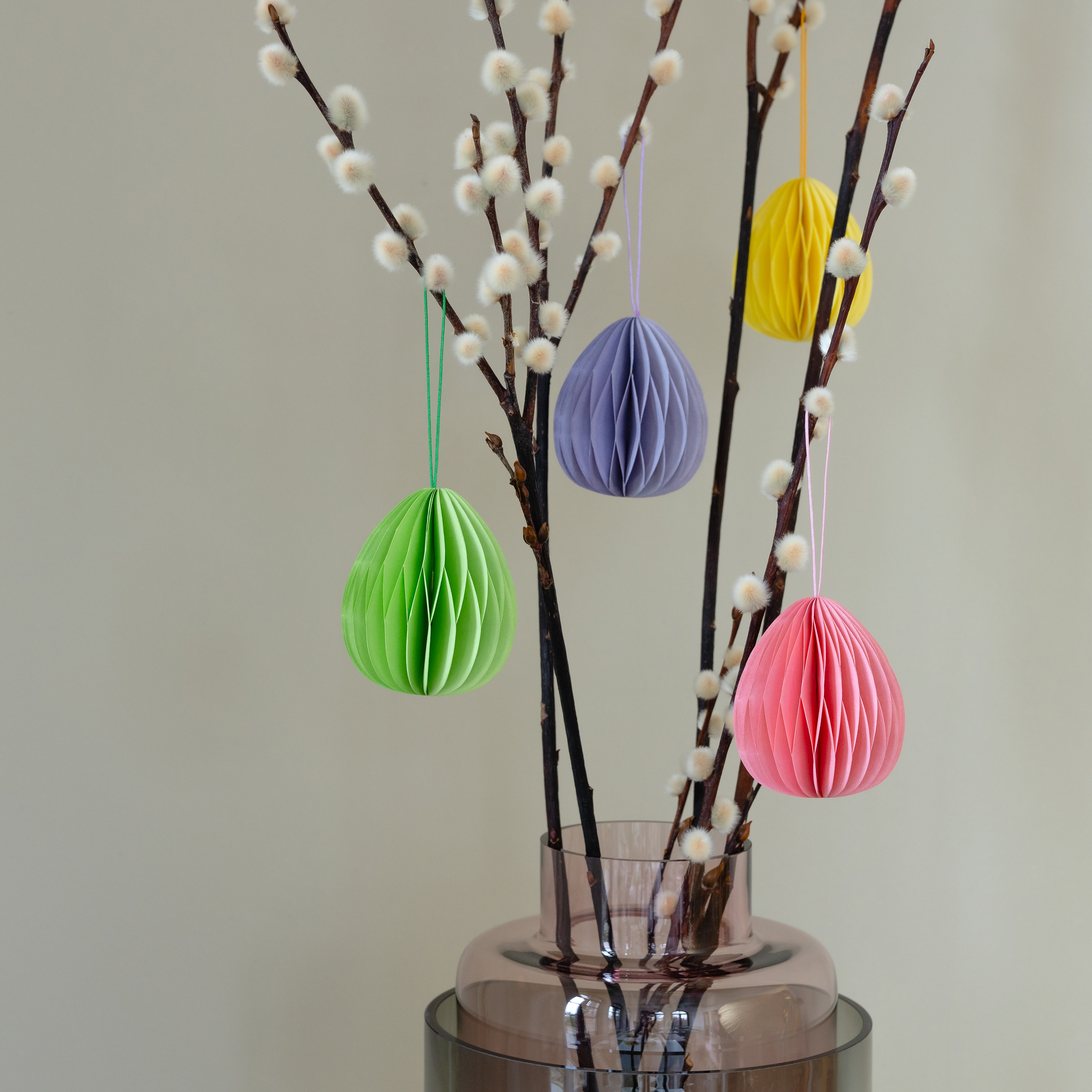 Delicados amentos de sauce en un jarrón con cuatro adornos de árbol ovalados y de colores, hechos de papel. Los colores son verde, lila, amarillo y rosa.