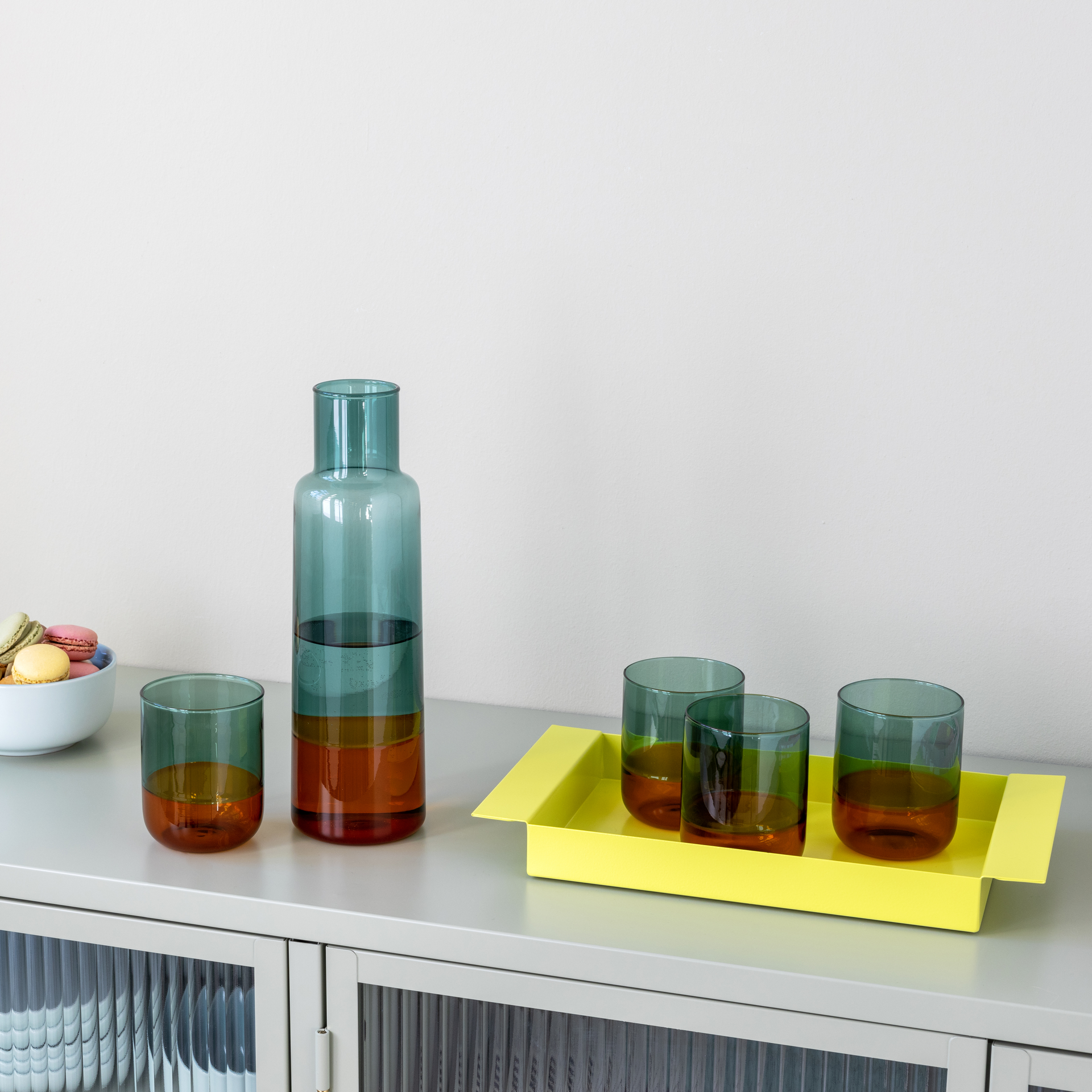 Two 'amber' glasses and a carafe on a tray. The drinking glasses are two-coloured: green and brown. A yellow tray and a bowl of macarons can also be seen.