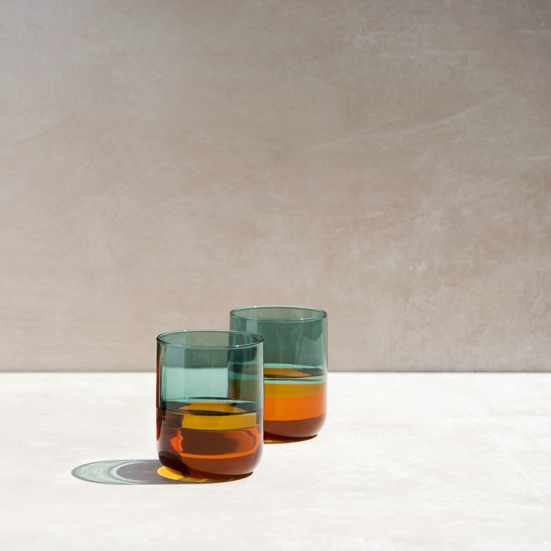 Two tall, slim glasses in an amber design stand on a light-coloured surface against a beige background. The glasses are partially filled with an amber-coloured liquid.