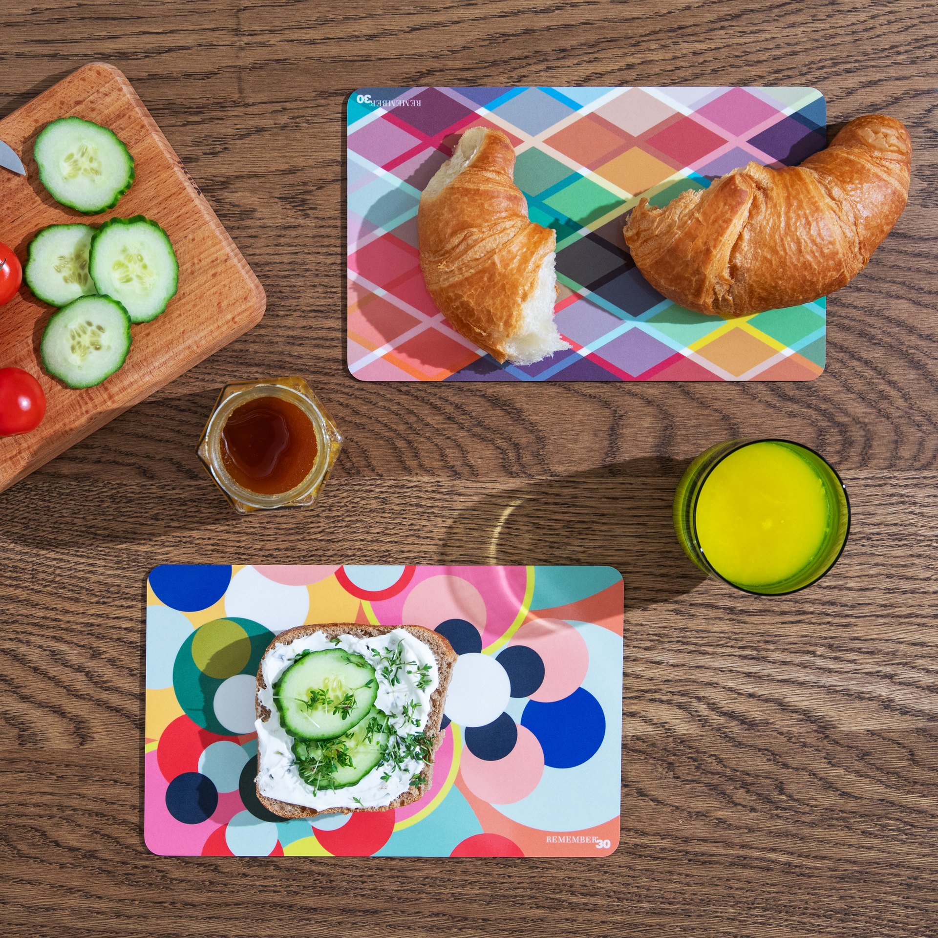 Frühstück auf einem Holztisch: Brot mit Gurke, Croissants, Honig, Saft, Gurken- und Tomatenscheiben auf einem Brett. Bunte Unterlagen unter den Speisen.