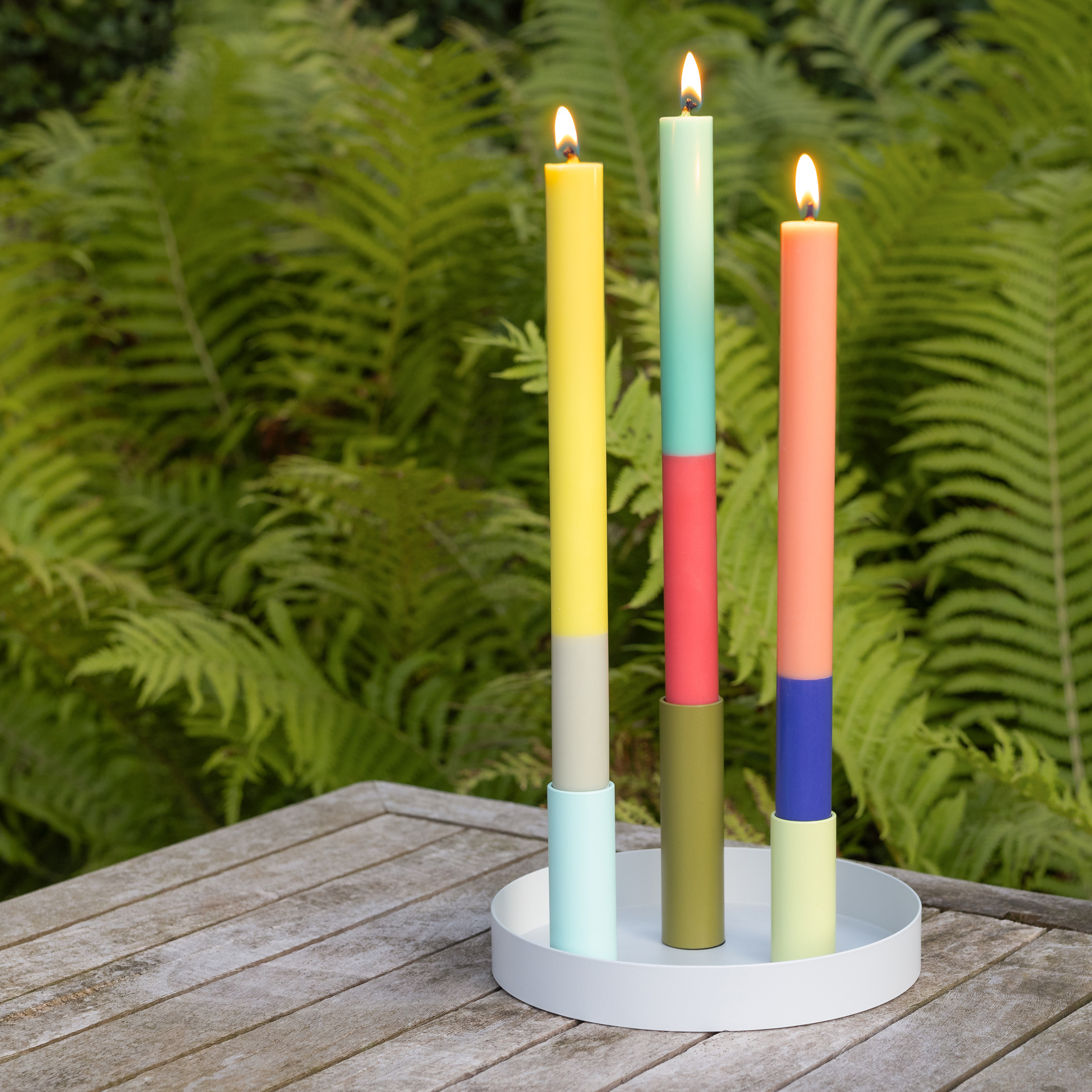Tres velas cilíndricas multicolores «Ibiza» encendidas sobre una bandeja redonda blanca, colocada sobre una superficie de madera, con un helecho verde al fondo.