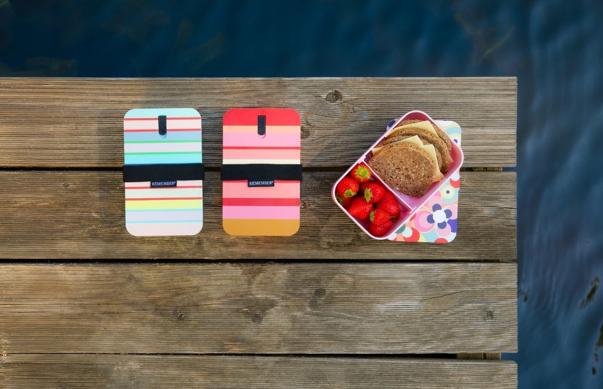 Zwei bunte, gestreifte Handyhüllen mit schwarzen Gummibändern und "REMEMBER"-Aufschrift liegen auf einem Holzsteg. Daneben eine offene Lunchbox mit frischen Erdbeeren und Vollkornbroten mit Käse. Im Hintergrund dunkles Wasser.