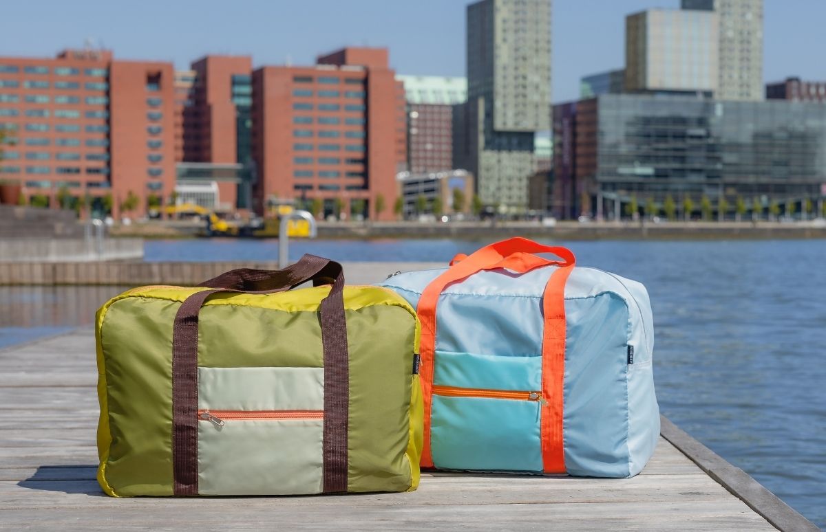 Zwei gefaltete Reisetaschen stehen auf einem Holzsteg am Wasser. Vorn olivgrün mit hellgrünen Details und braunen Henkeln, hinten hellblau mit aquamarinfarbenen Details und leuchtend orangen Henkeln. Moderne Gebäude am Ufer.