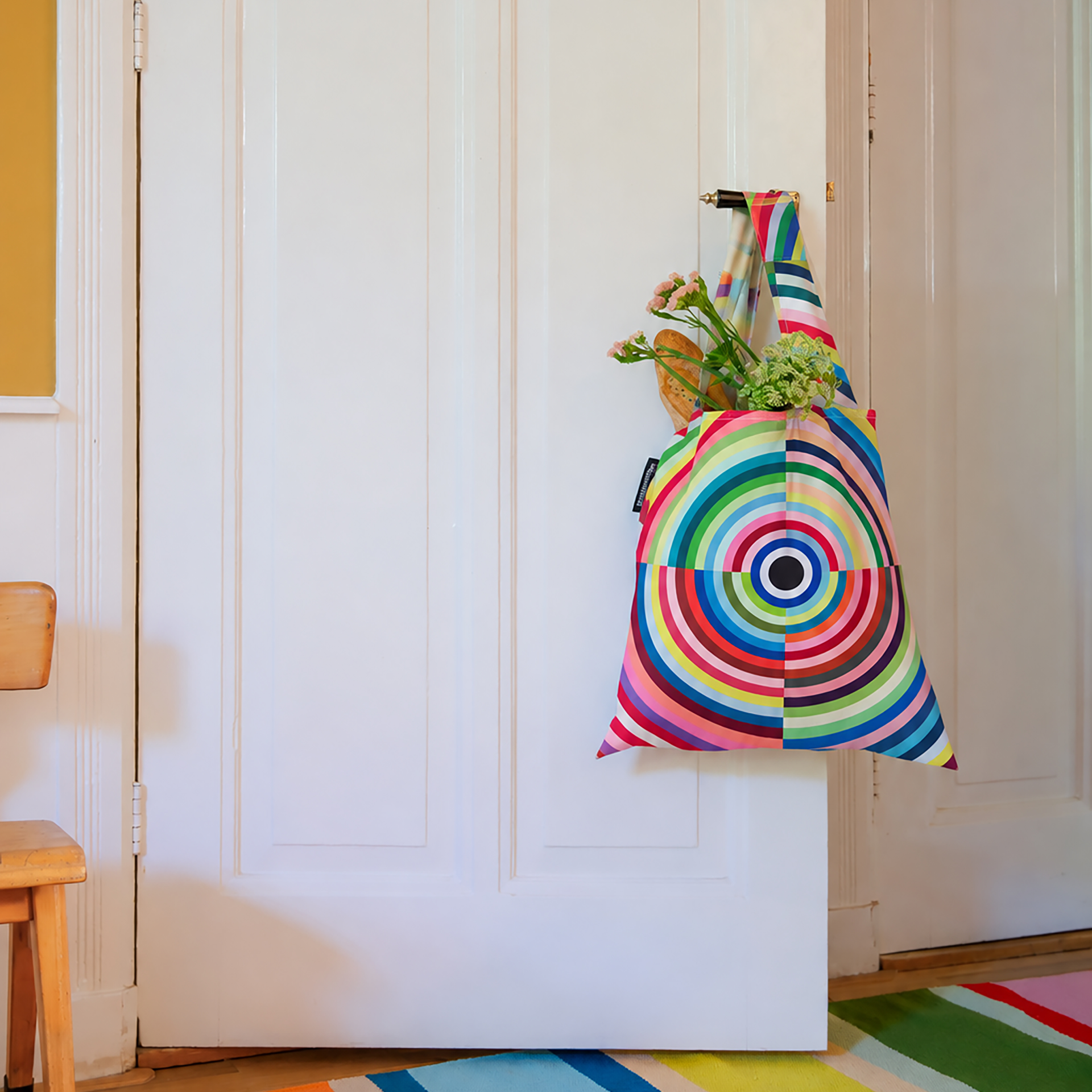 Faltbare Tragetasche mit Mandala-Muster aus konzentrischen Ringen in Rot, Blau, Grün und Gelb hängt an weißem Türgriff. In der Tasche sind Baguette und frische Blumen. Bunter Teppich auf Holzboden.