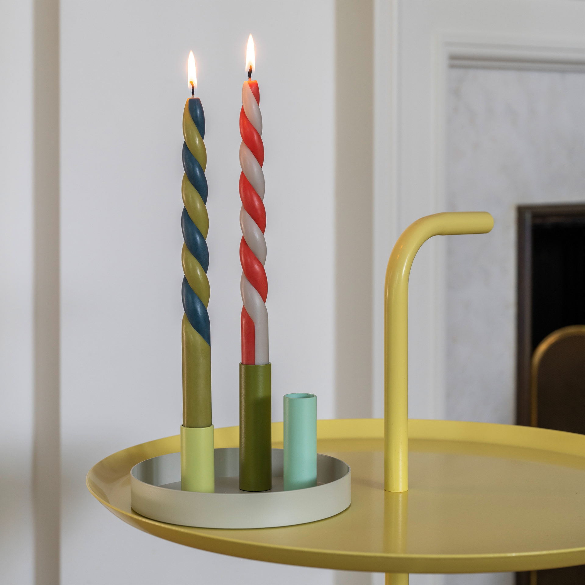 Dos velas cilíndricas de colores «Vienna» encendidas sobre una mesa redonda amarilla con candelabros a juego. Las velas son torcidas.