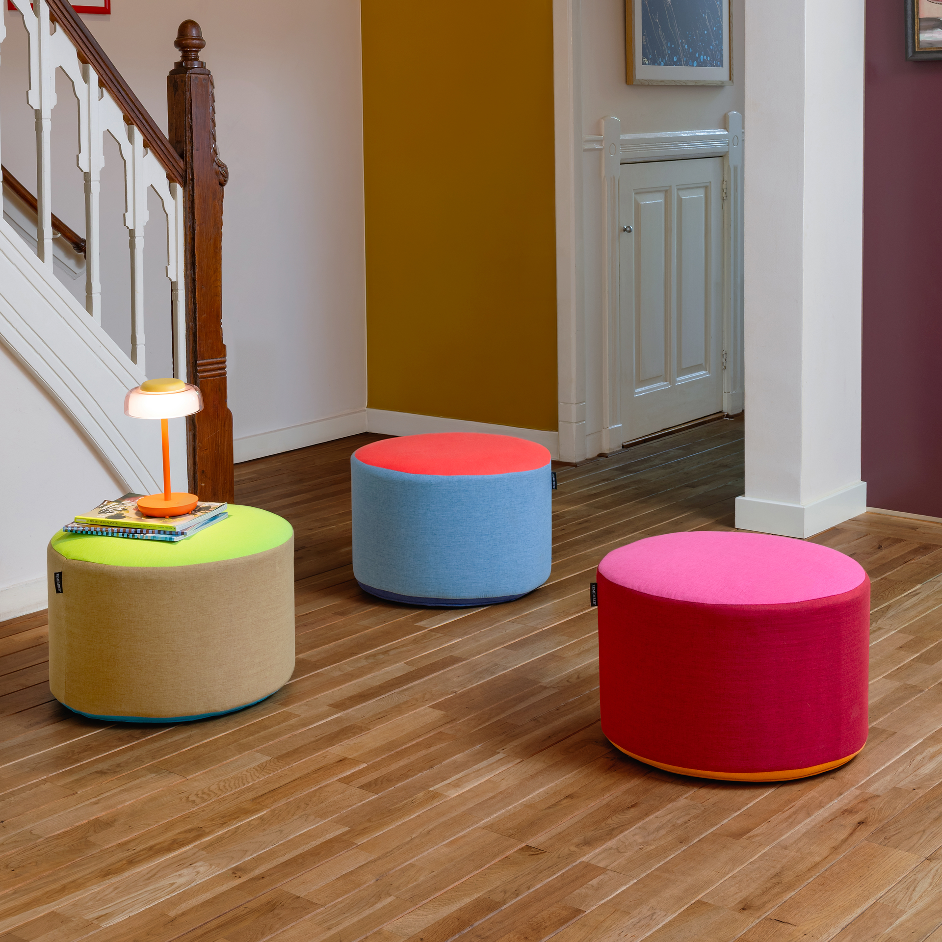 Pouf redondo com revestimento têxtil tricolor: parte superior em verde vivo, corpo em tom areia claro e parte inferior com uma faixa estreita em turquesa. Pequena etiqueta preta « REMEMBER » na lateral. Convida a sentar-se e a relaxar.