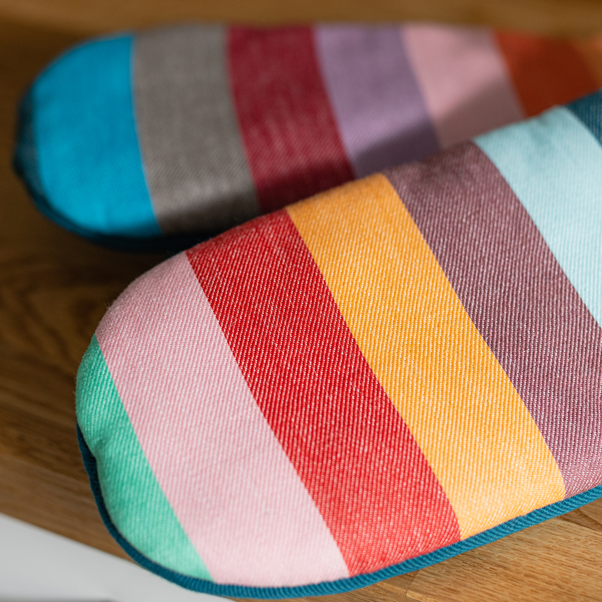 Primo piano di due guanti da forno in un set. Strisce colorate in verde, rosa, rosso, giallo, marrone, turchese, grigio e viola. I guanti sono appoggiati l'uno sull'altro su una superficie di legno.