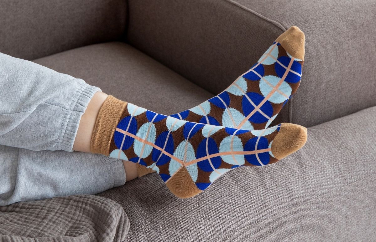Ein Paar Füße in gemusterten Socken ruht bequem auf einer grauen Couch. Die Socken zeigen ein Argyle-ähnliches Muster aus braunen Rauten, blauen und hellblauen Kreisen, durchzogen von feinen rosa Linien. Die Person trägt graue Freizeithosen.