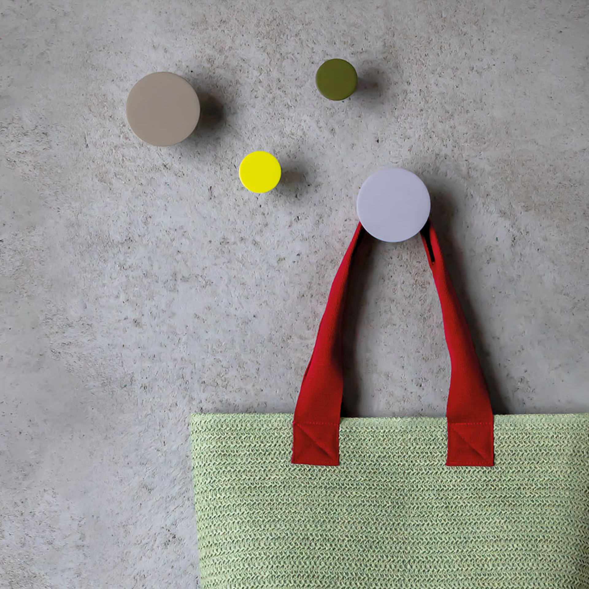 Four wall hooks in beige, green, yellow and purple on a wall, with a light green bag with red handles hanging from one hook.