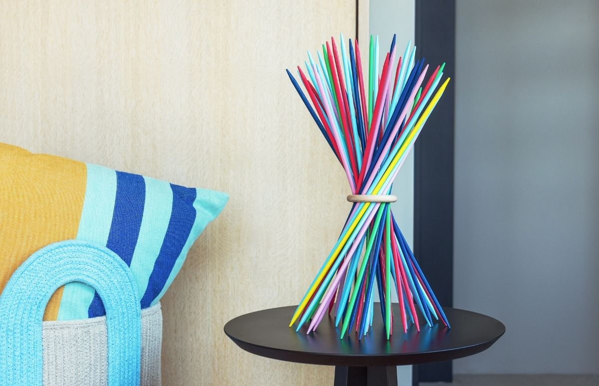Bunter Strauß Mikado-Stäbchen, mittig mit Holzring gebunden, fächert sich auf dunklem Beistelltisch auf. Links ein gestreiftes Kissen (gelb/blau) auf dem Sofa, daneben ein heller Beutel mit blauem Henkel. Helle Holzwand und dunkelgraue Wand.