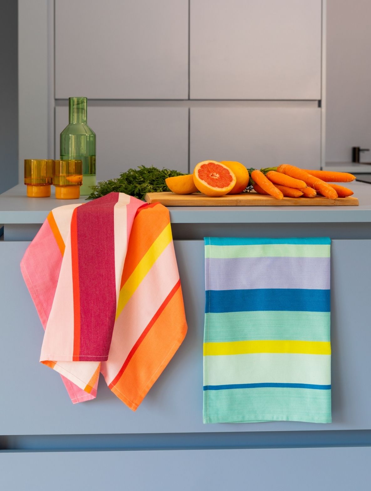 Moderne Küche mit blauer Arbeitsplatte. Darauf: grüne Flasche, zwei gelbe Gläser und Holzplatte mit halbierter Grapefruit, Karotten und frischen Kräutern. Zwei bunte, gestreifte Tücher in Rosa-Orange-Gelb und Blau-Grün hängen über die Kante.