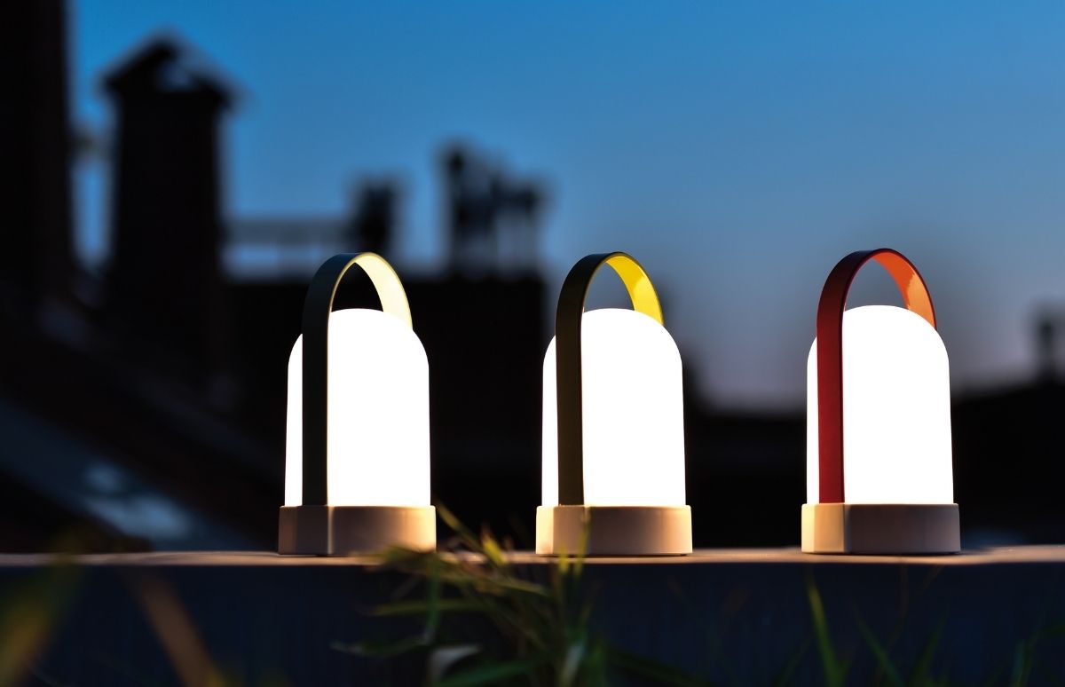 Drei leuchtende, moderne tragbare Lampen stehen nebeneinander auf dunkler Oberfläche im Freien bei Dämmerung. Weiße Leuchtkörper, farbige Griffe in Dunkelgrün, Gelb und Rot. Unscharfer blauer Nachthimmel, Silhouetten.