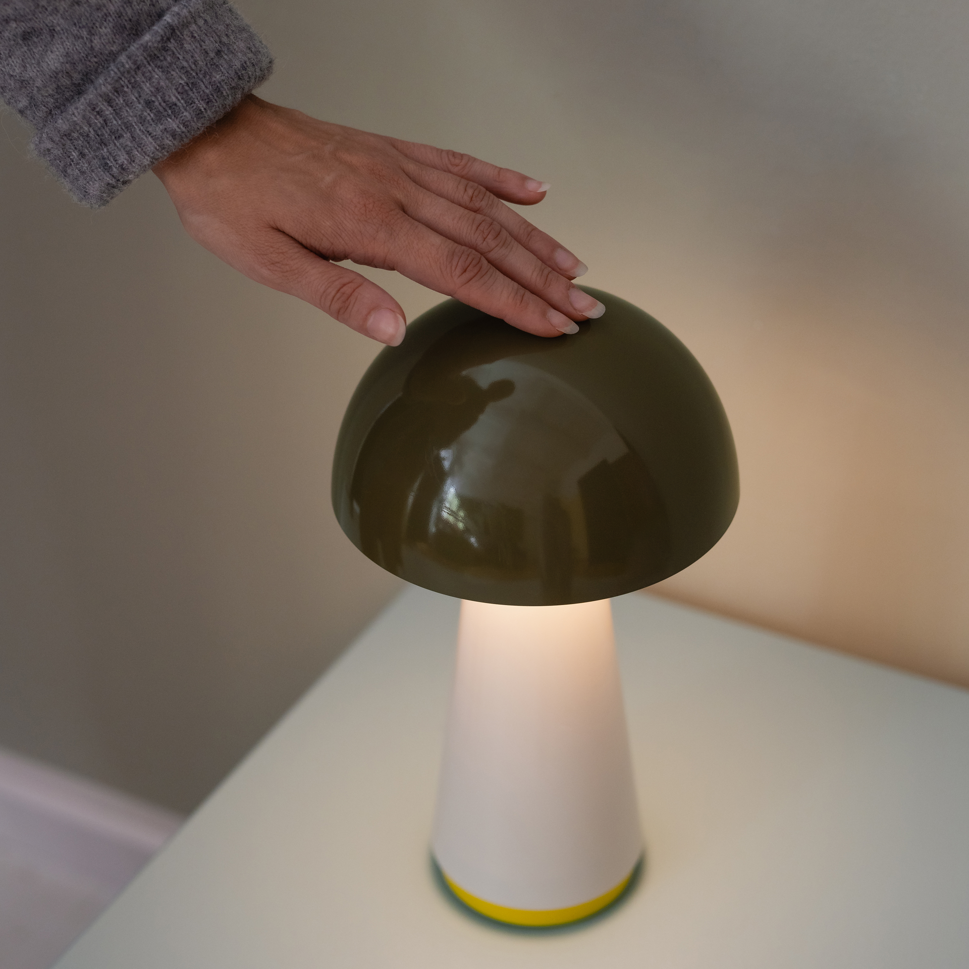 Primer plano de la lámpara de mesa Bob en verde oliva. Una mano toca la pantalla. La luz de la lámpara es de color blanco cálido y brilla con discreción.