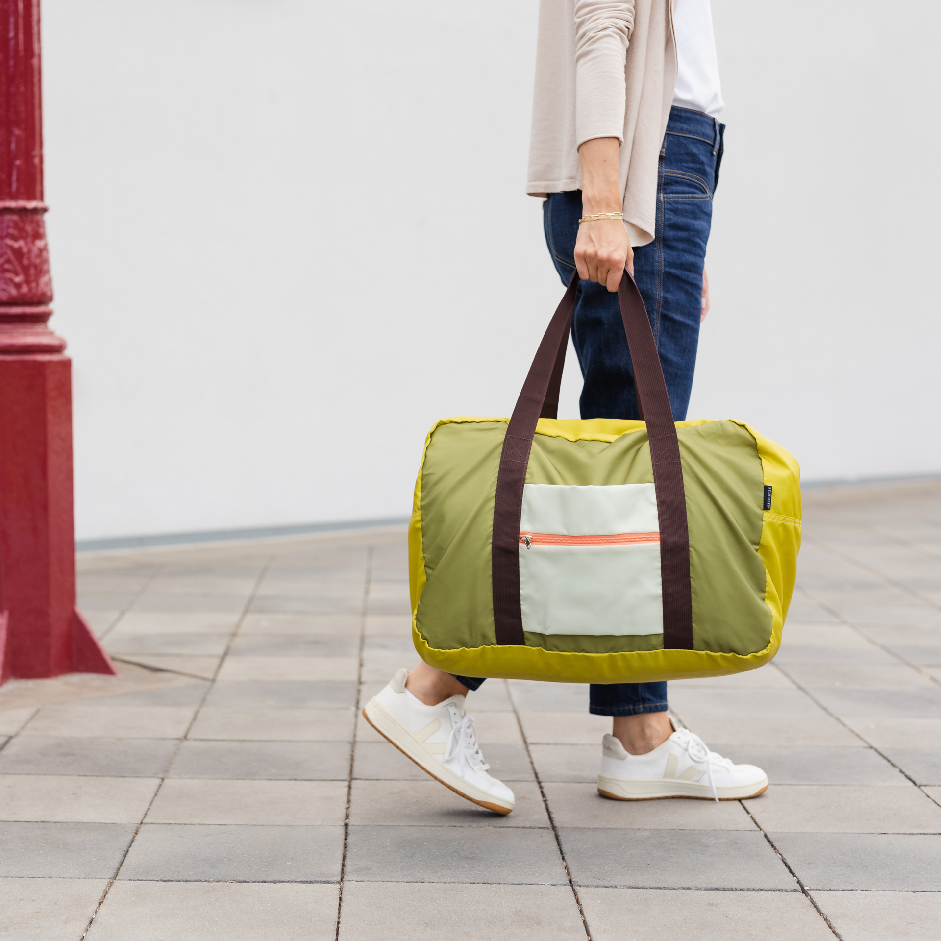 Una persona lleva la bolsa de viaje verde «Liz» con asas marrones. La bolsa tiene un bolsillo exterior claro con cremallera. Lleva vaqueros y zapatillas blancas.