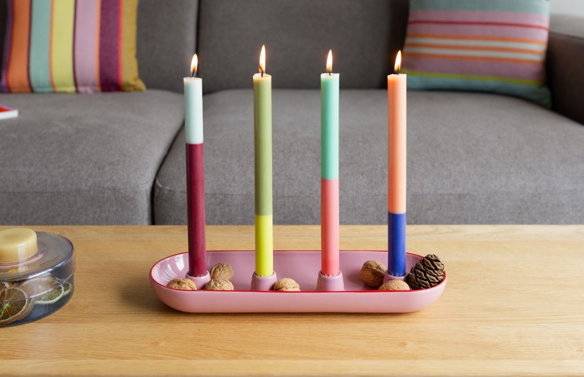 4 kleurrijke kaarsen in een roze kaarsenhouder op een houten tafel; walnoten en dennenappels. Kleuren: blauw/rood, groen/geel, mint/roze, perzik/blauw. Grijze bank met gestreept kussen op de achtergrond.