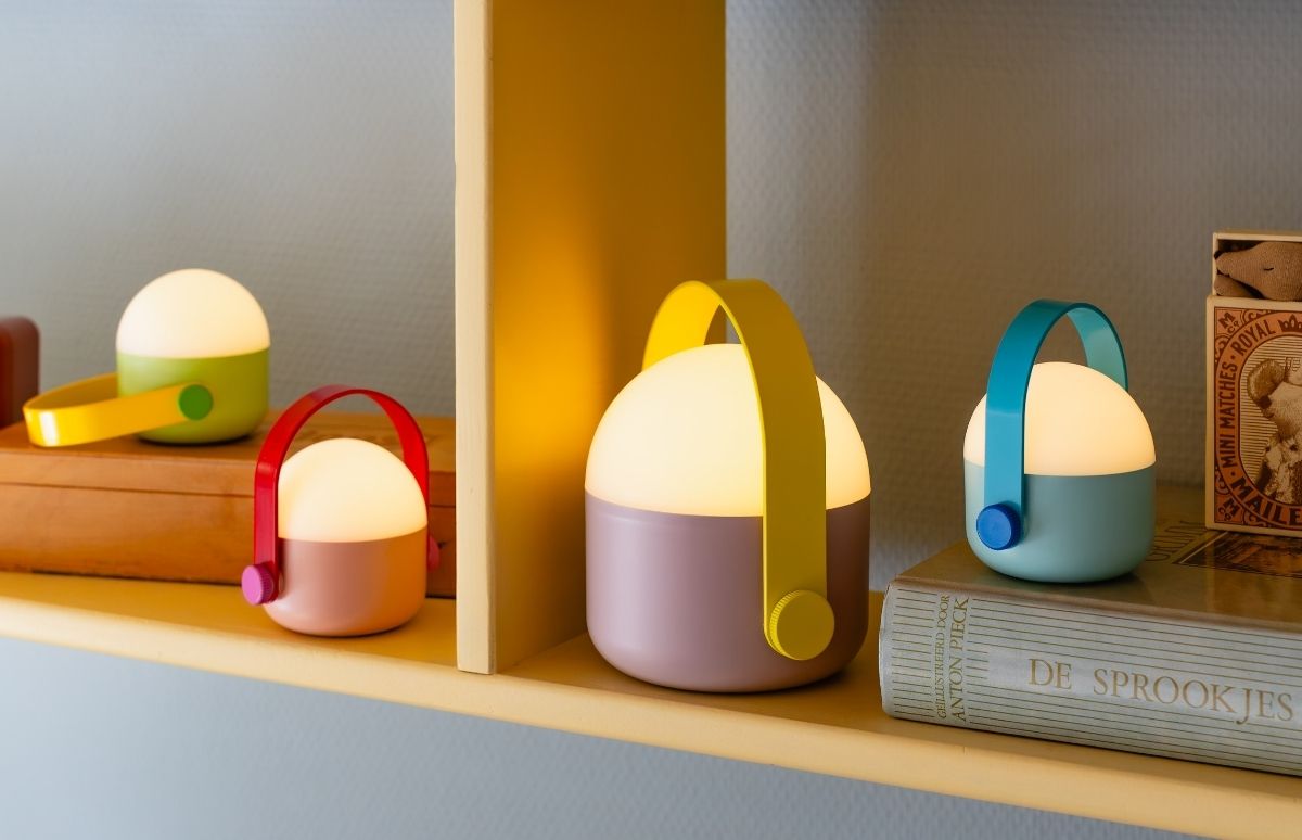 Vier leuchtende, tragbare LED-Lampen in Pastellfarben auf Holzregalen. Jede Lampe mit weißer Kuppel, farbigem Sockel und passendem Tragegriff. Farbkombinationen: Grün/Gelb, Rosa/Rot, Mauve/Gelb, Blaugrün/Blau.